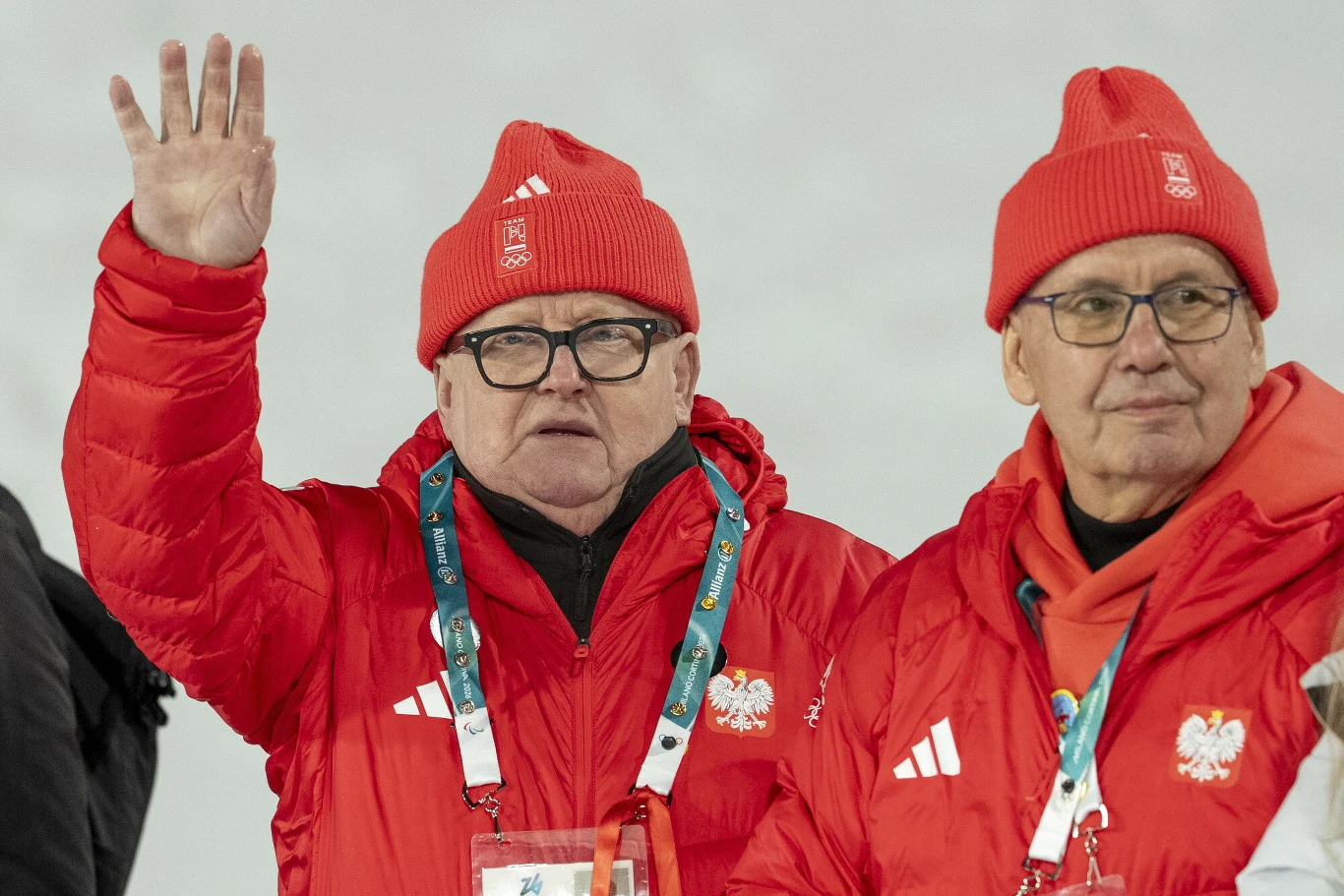 Dwóch mężczyzn w czerwonych zimowych kurtkach i czapkach z biało-czerwonymi emblematami, jeden z nich w okularach podnosi rękę w geście pozdrowienia; obaj mają na szyjach identyfikatory i niebieskie sznury, a w tle widoczna jest jednolita, jasna przest...