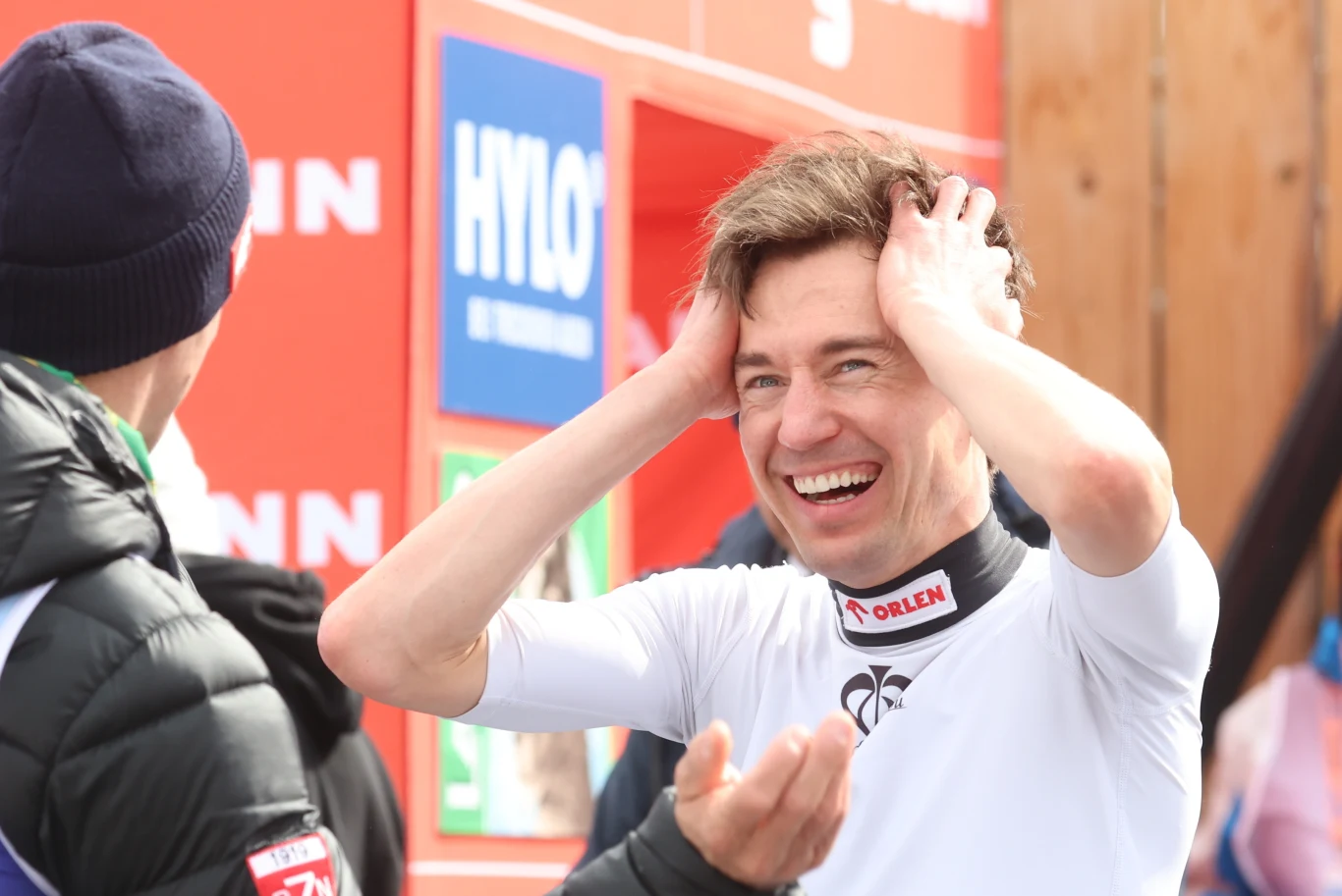 sportowiec w białym stroju z uśmiechem na twarzy trzyma się za głowę, rozmawiając z osobą w czarnej kurtce; w tle czerwone tablice z widocznymi logotypami sponsorskimi