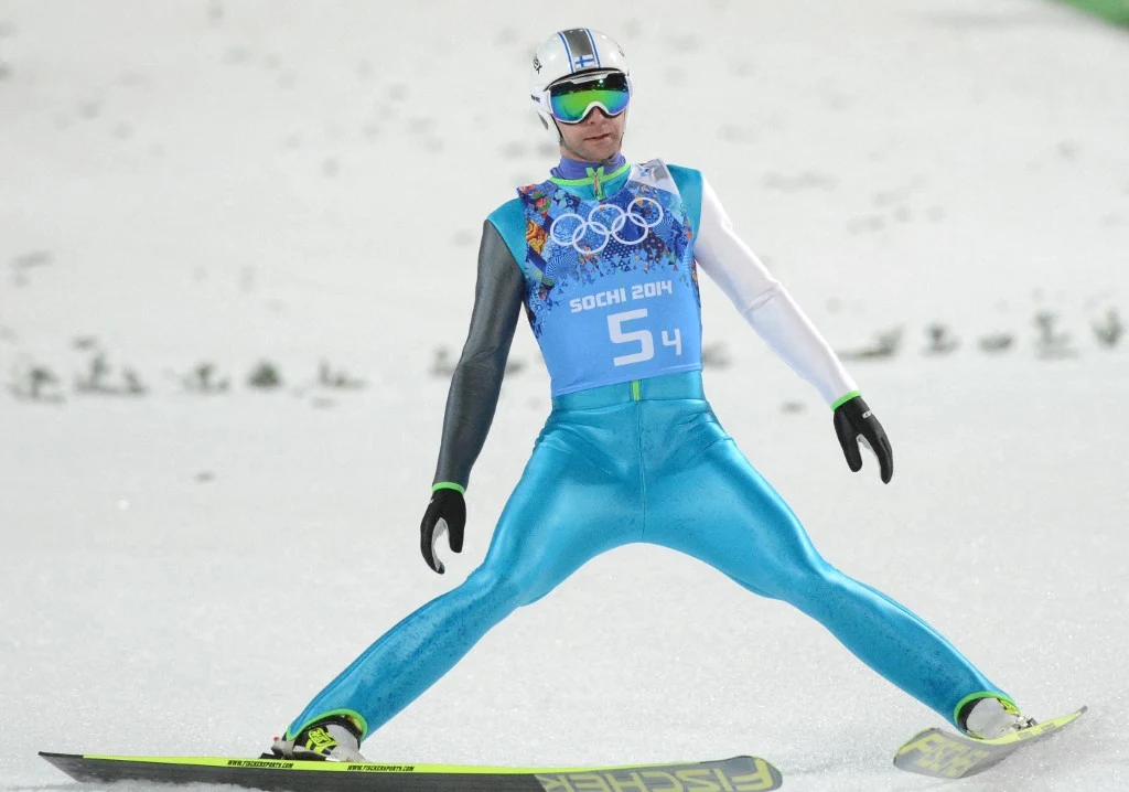 Skoczek narciarski w lądowaniu na śniegu, ubrany w niebieski kombinezon, z numerem 54, uczestnik zimowych igrzysk olimpijskich Soczi 2014, w goglach i kasku ochronnym, szeroko rozstawione narty podczas fazy dojazdu po skoku.