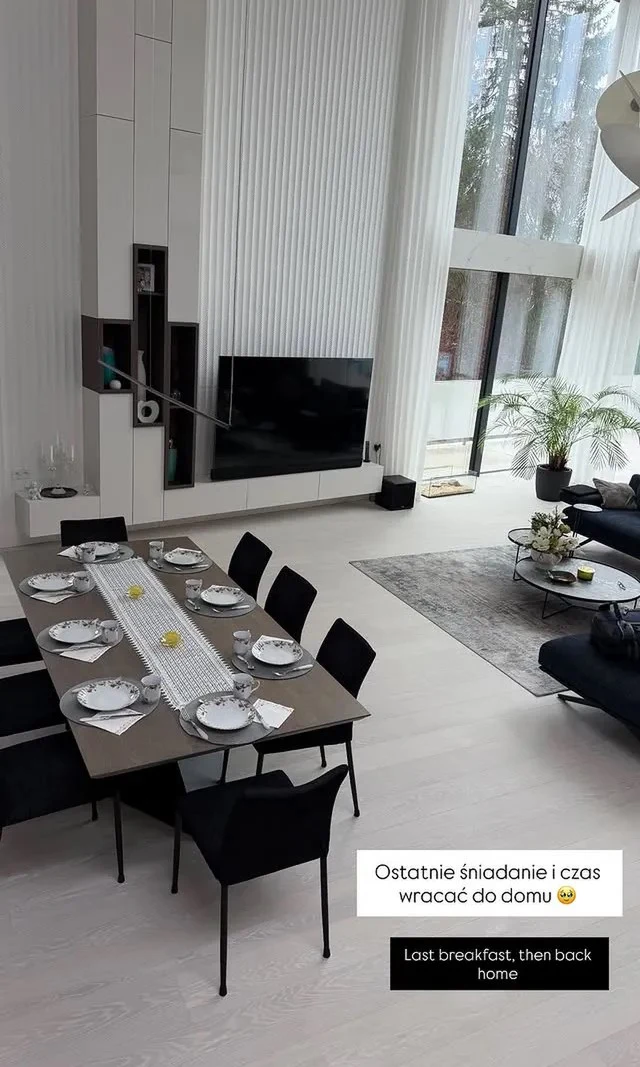 nowocześnie urządzony salon z dużymi oknami, jadalnią przygotowaną do śniadania dla ośmiu osób, jasną podłogą i czarną kanapą, w tle wysoka ściana z telewizorem oraz dekoracyjną rośliną przy oknie