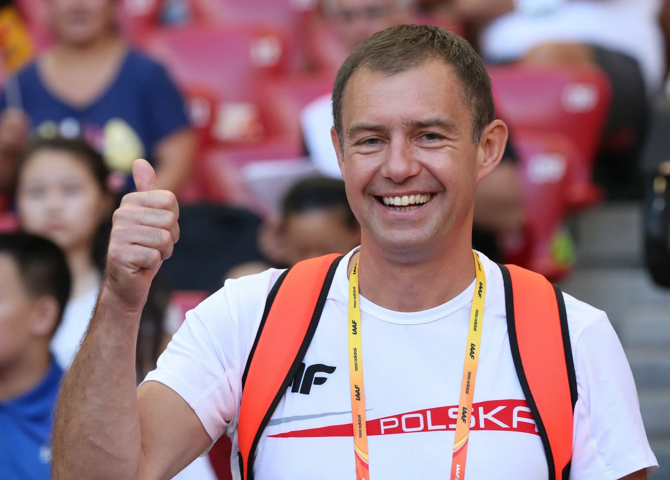 uśmiechnięty mężczyzna w białej koszulce z napisem 'Polska', z pomarańczowym plecakiem na ramionach i akredytacją na szyi, pokazuje uniesiony kciuk w geście aprobaty, siedząc na stadionowych trybunach