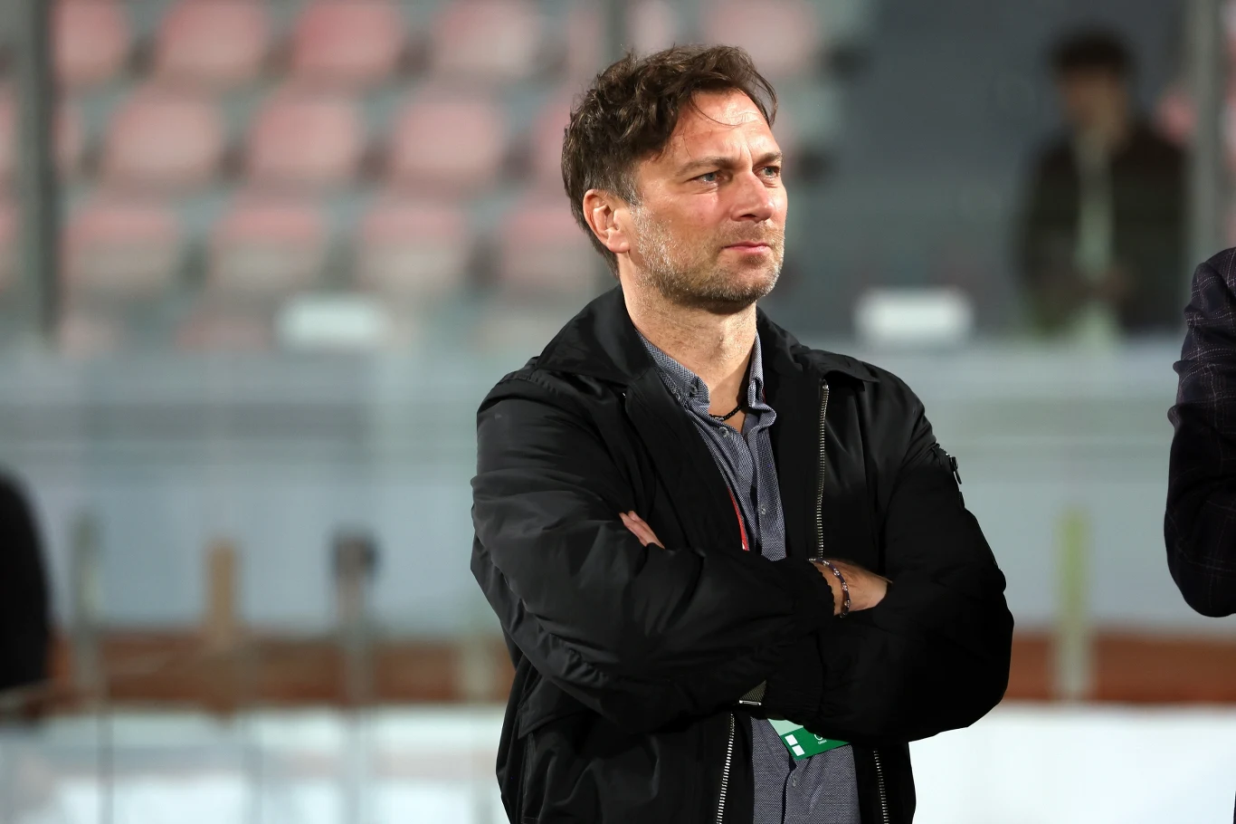 mężczyzna w średnim wieku z krótkimi włosami stoi z założonymi rękami w sportowym otoczeniu, ubrany w ciemną kurtkę, w tle rozmyte siedzenia stadionowe