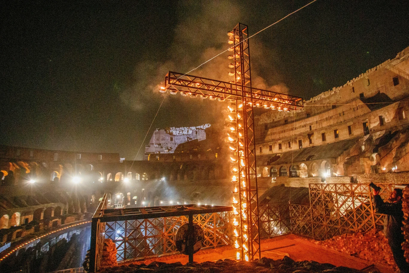 duży podświetlony krzyż z płonącymi światłami ustawiony wewnątrz antycznego amfiteatru, wokół którego widać nocną scenerię i częściowo oświetlone kamienne mury Koloseum