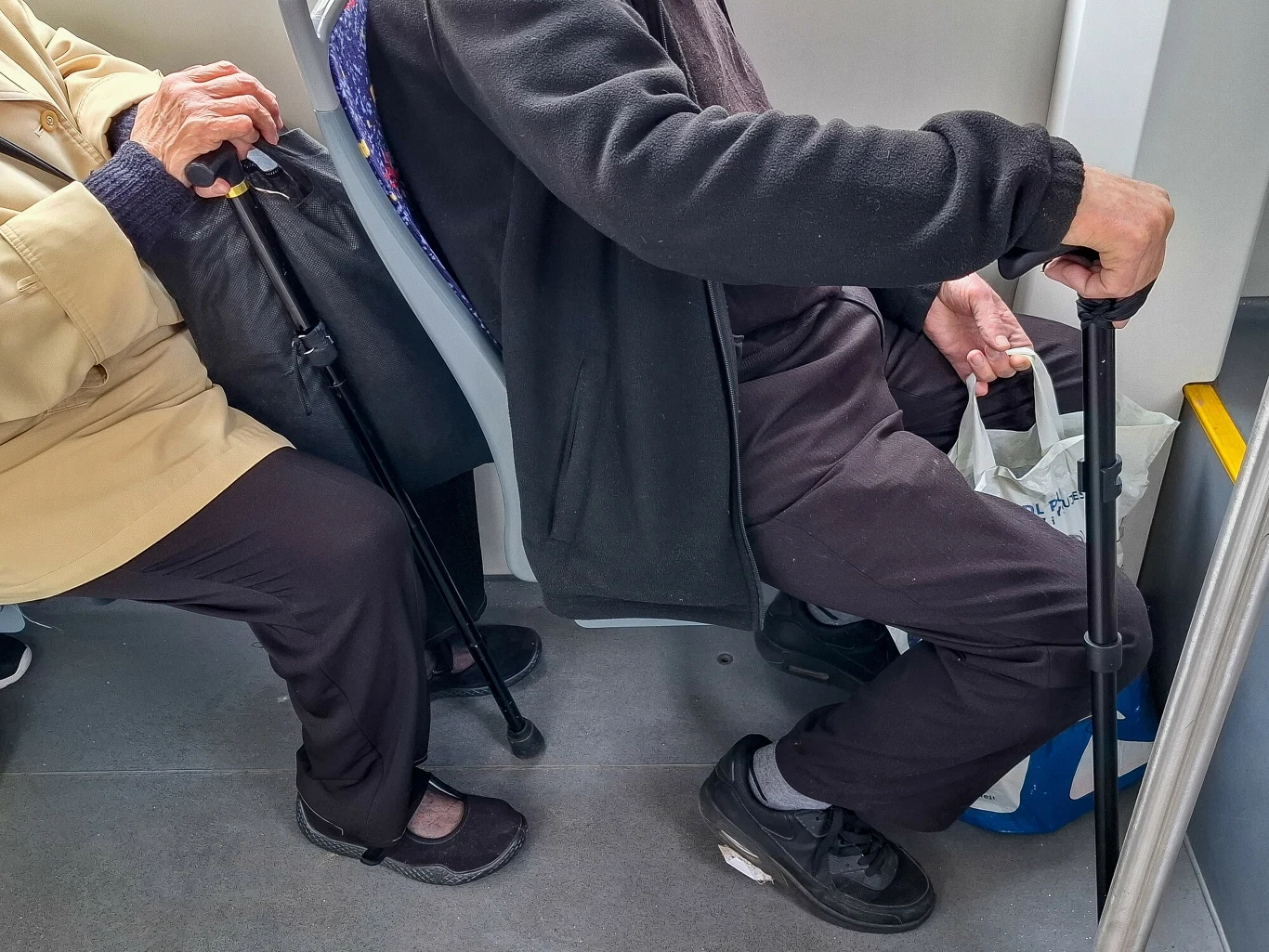 dwie starsze osoby siedzące obok siebie w środku komunikacji miejskiej, każda trzyma laskę wspierającą, jedna osoba ma torbę na zakupy na podłodze