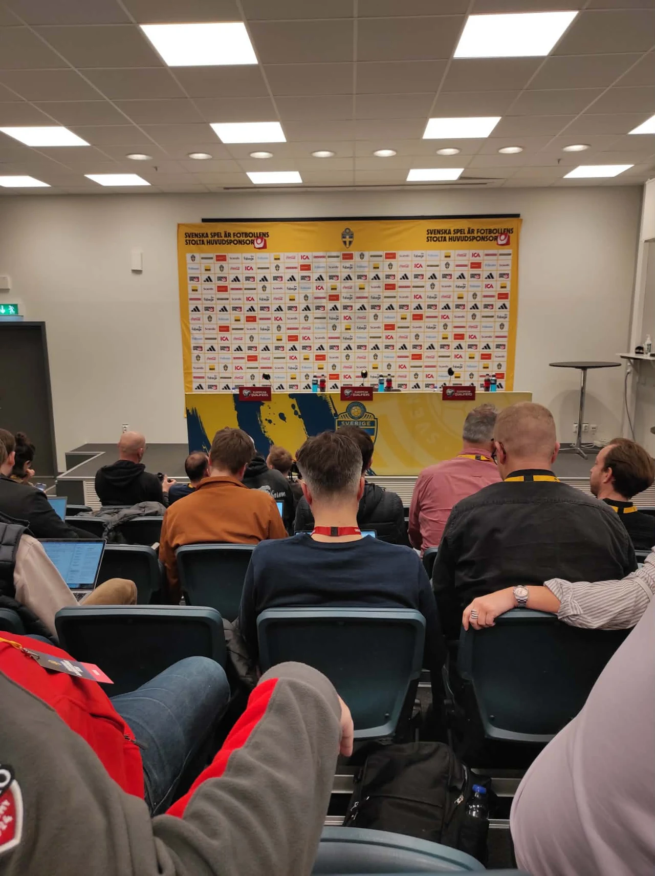 sala konferencyjna z uczestnikami siedzącymi tyłem do kamery, na wprost duża ścianka sponsora z wieloma logotypami, z przodu ustawione dwa stoły z mikrofonami gotowe do konferencji prasowej