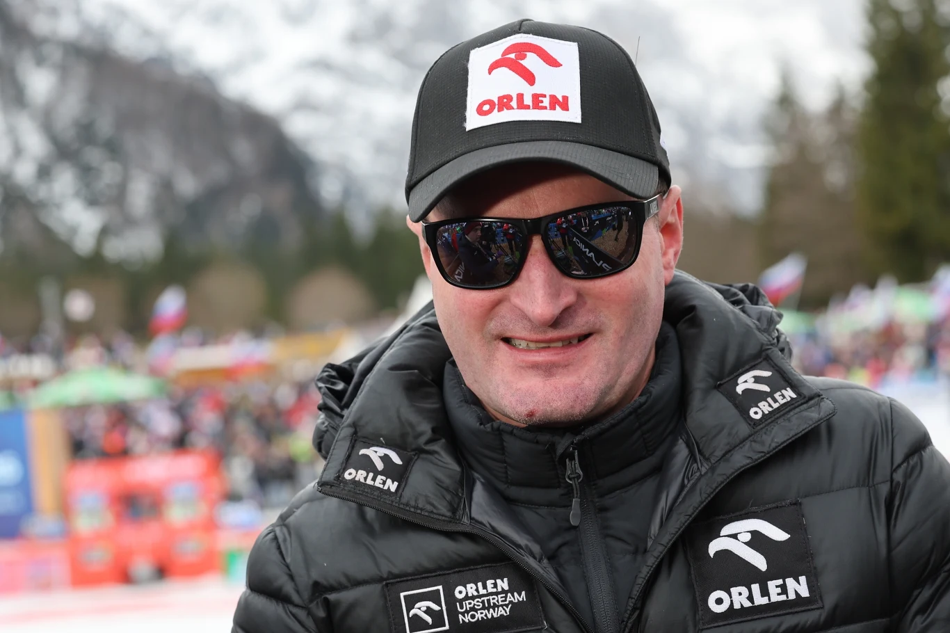 Mężczyzna w ciemnych okularach i czapce z logo ORLEN, ubrany w czarną sportową kurtkę z tym samym logotypem, znajduje się na tle gór i zgromadzonych kibiców, co sugeruje zimową imprezę sportową.