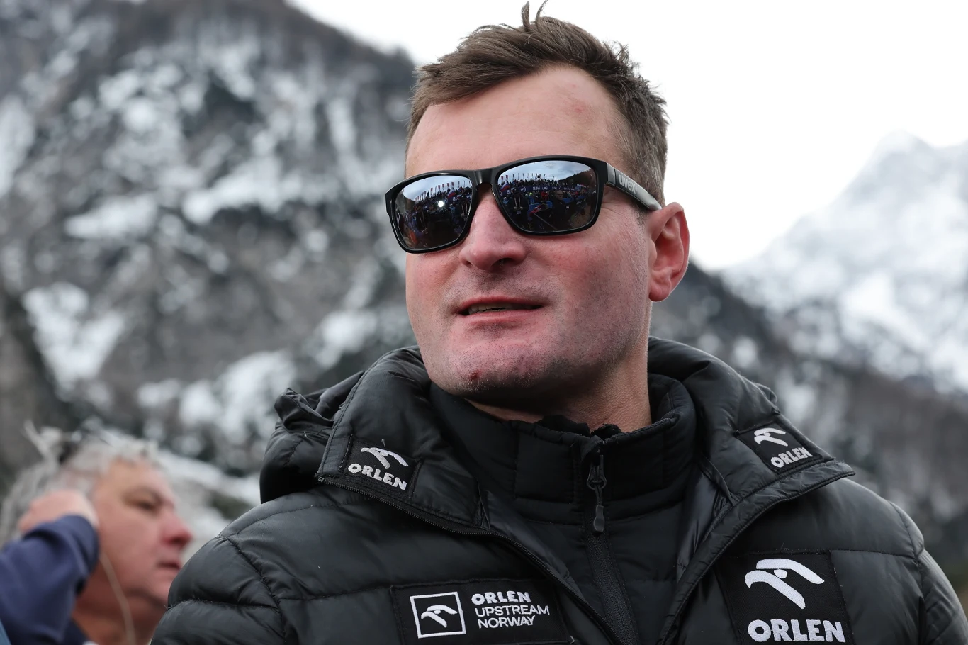 Mężczyzna w ciemnych okularach przeciwsłonecznych i kurtce zimowej z logo Orlen na tle ośnieżonych gór.