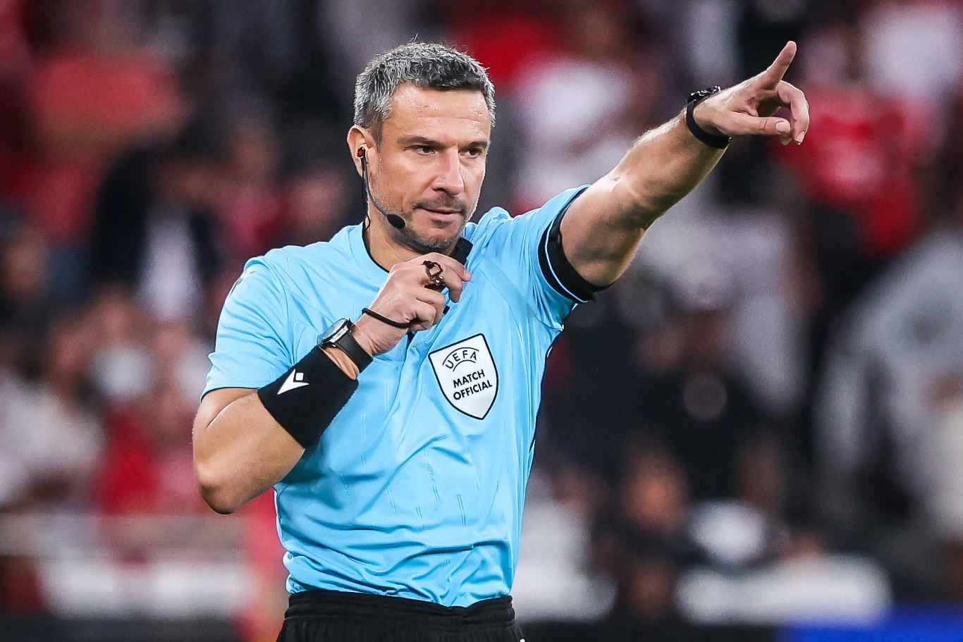 Sędzia piłkarski w niebieskim stroju z odznaką UEFA, używający gwizdka i wskazujący ręką kierunek podczas meczu.