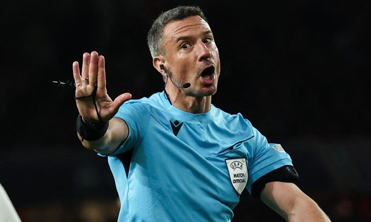 Sędzia piłkarski w niebieskim stroju z uniesioną ręką w geście zatrzymania, z mikrofonem przy twarzy, na ciemnym tle stadionu.