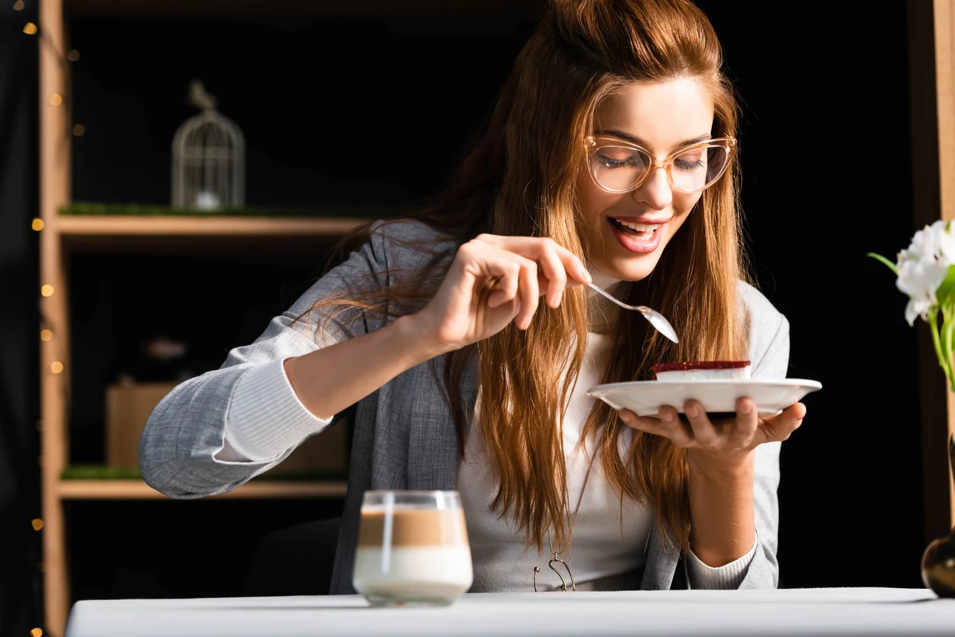 uśmiechnięta kobieta w okularach trzyma talerz z deserem i je go łyżeczką, na pierwszym planie filiżanka kawy oraz wazonik z białymi kwiatami