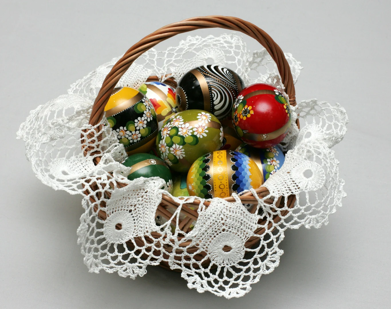 Koszyk z wikliny ozdobiony białą, szydełkową serwetką, wypełniony kolorowymi, ręcznie malowanymi pisankami o różnych wzorach i motywach, głównie kwiatowych i geometrycznych.