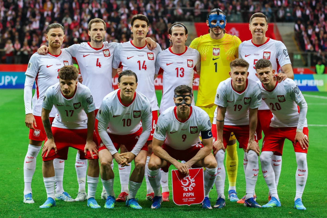 jedenastu piłkarzy reprezentacji Polski w białych strojach drużynowych pozuje na stadionie do wspólnego zdjęcia przed meczem, w tle wypełnione trybuny z kibicami i biało-czerwonymi flagami, jeden z zawodników nosi czarną maskę ochronną na twarzy, a bra...