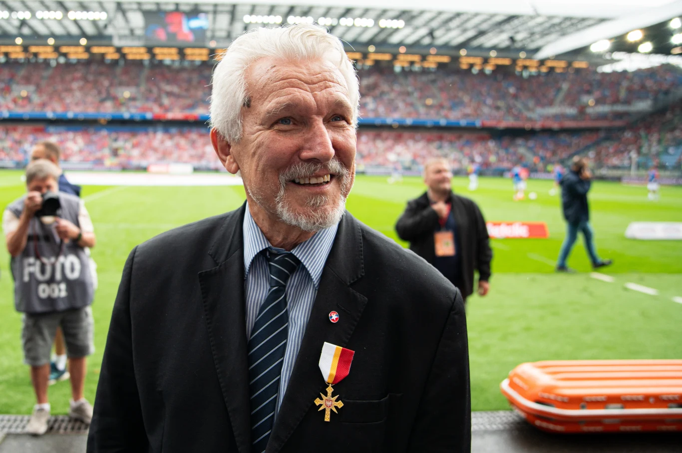 starszy mężczyzna w garniturze i krawacie z przypiętym odznaczeniem na piersi stoi na tle stadionu piłkarskiego, wokół wielu ludzi oraz fotograf z aparatem; w tle trybuny wypełnione kibicami