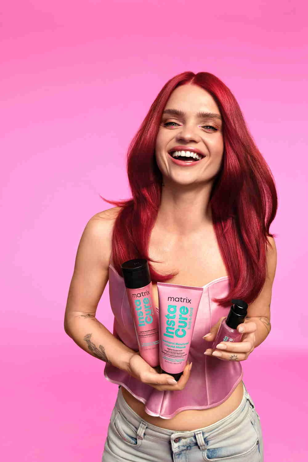 Margaret, znana z odważnych metamorfoz, przyznaje, że włosy muszą nadążać za tempem jej życia uśmiechnięta kobieta z długimi, intensywnie czerwonymi włosami trzyma trzy produkty do pielęgnacji włosów marki Matrix Insta Cure, ubrana w jasnoróżowy gorset i jasne jeansy na tle różowego tła