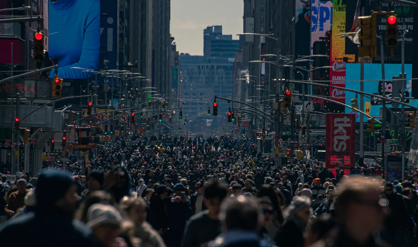 Tłum ludzi wypełnia szeroką ulicę w centrum miasta, wokół liczne światła sygnalizacji świetlnej, wieżowce oraz kolorowe reklamy na budynkach.