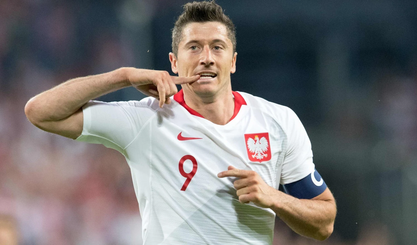 Robert Lewandowski Piłkarz w białej koszulce z orłem i numerem 9 wykonuje charakterystyczny gest dłonią przy ustach, na lewej ręce opaska kapitana.