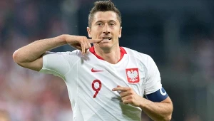 Piłkarz w białej koszulce z orłem i numerem 9 wykonuje charakterystyczny gest dłonią przy ustach, na lewej ręce opaska kapitana.