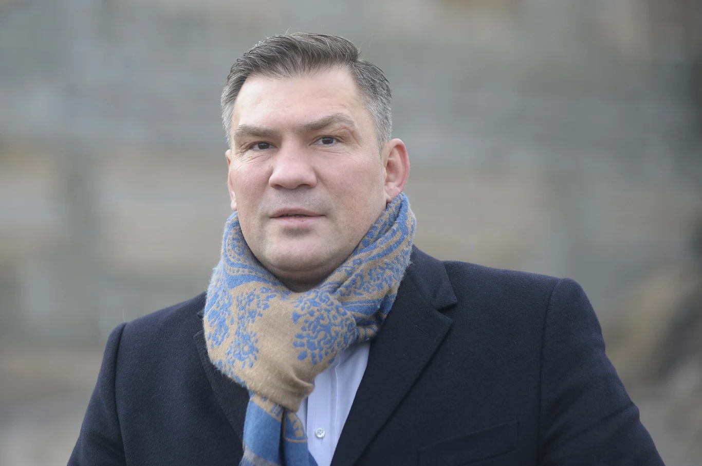Dariusz Michalczewski mężczyzna w ciemnym płaszczu i niebiesko-beżowym szaliku stoi na tle rozmytego, kamiennego muru, patrząc prosto przed siebie z poważnym wyrazem twarzy