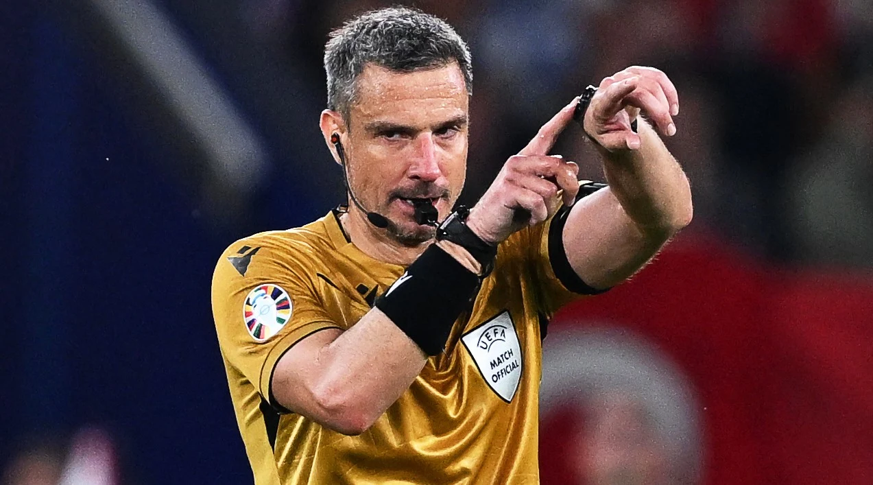 sędzia piłkarski w złotej koszulce sygnalizujący decyzję podczas meczu, używający zestawu słuchawkowego i zegarka VAR, w tle rozmyte trybuny stadionu