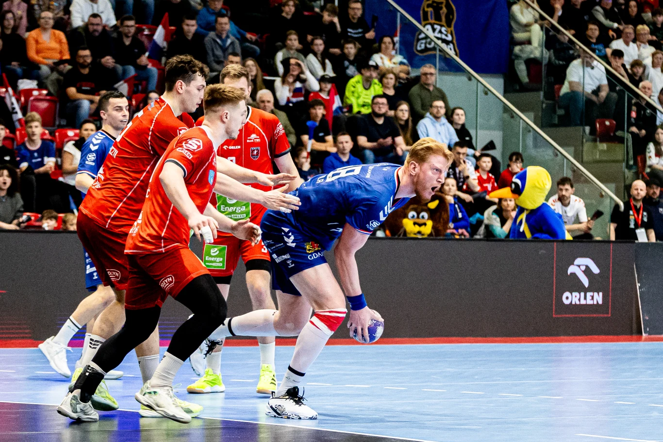 dwóch zawodników drużyny w czerwonych strojach próbuje powstrzymać szczypiornistę w niebieskim stroju z piłką, dynamiczna akcja podczas meczu piłki ręcznej, trybuny wypełnione kibicami