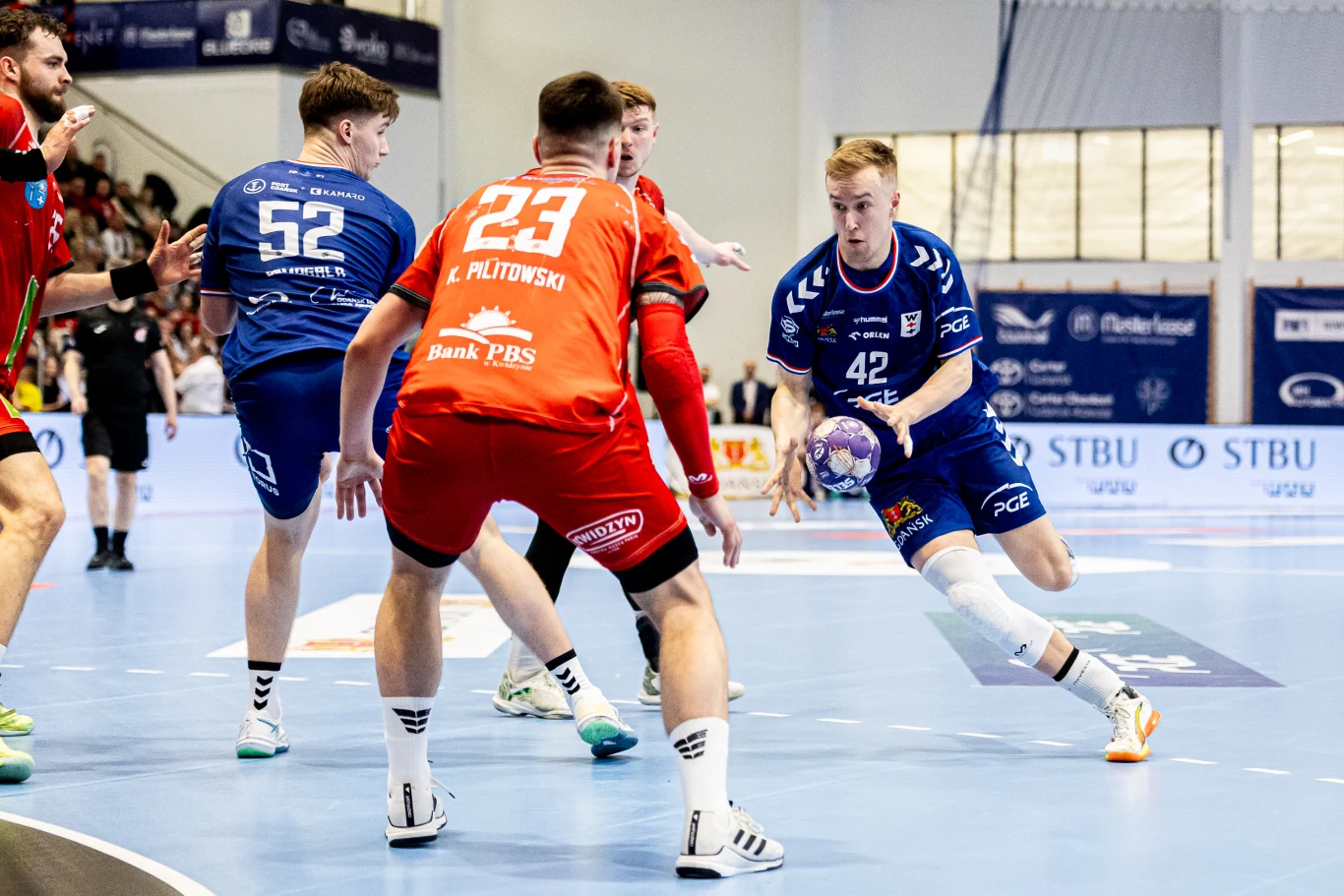 zawodnik w niebieskiej koszulce z piłką w rękach przygotowuje się do ataku, ścigany przez obrońcę w czerwonym stroju, na hali sportowej w trakcie meczu piłki ręcznej