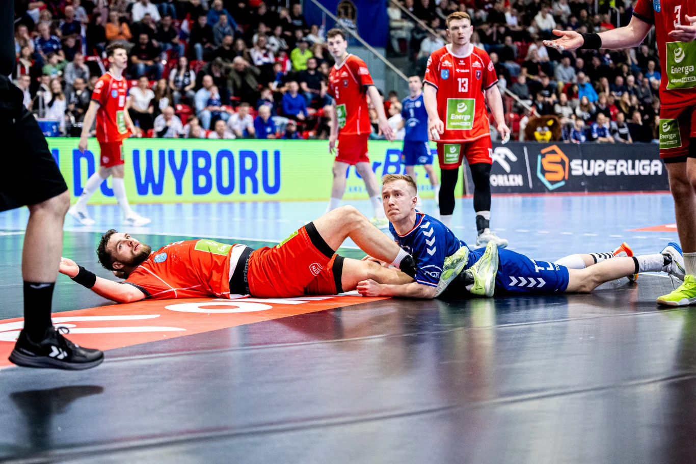 dwóch zawodników leżących na parkiecie podczas meczu piłki ręcznej, jeden ubrany w czerwony strój, drugi w niebieski, wokół nich kilku stojących graczy, w tle trybuny pełne kibiców oraz widoczne reklamy Superligi