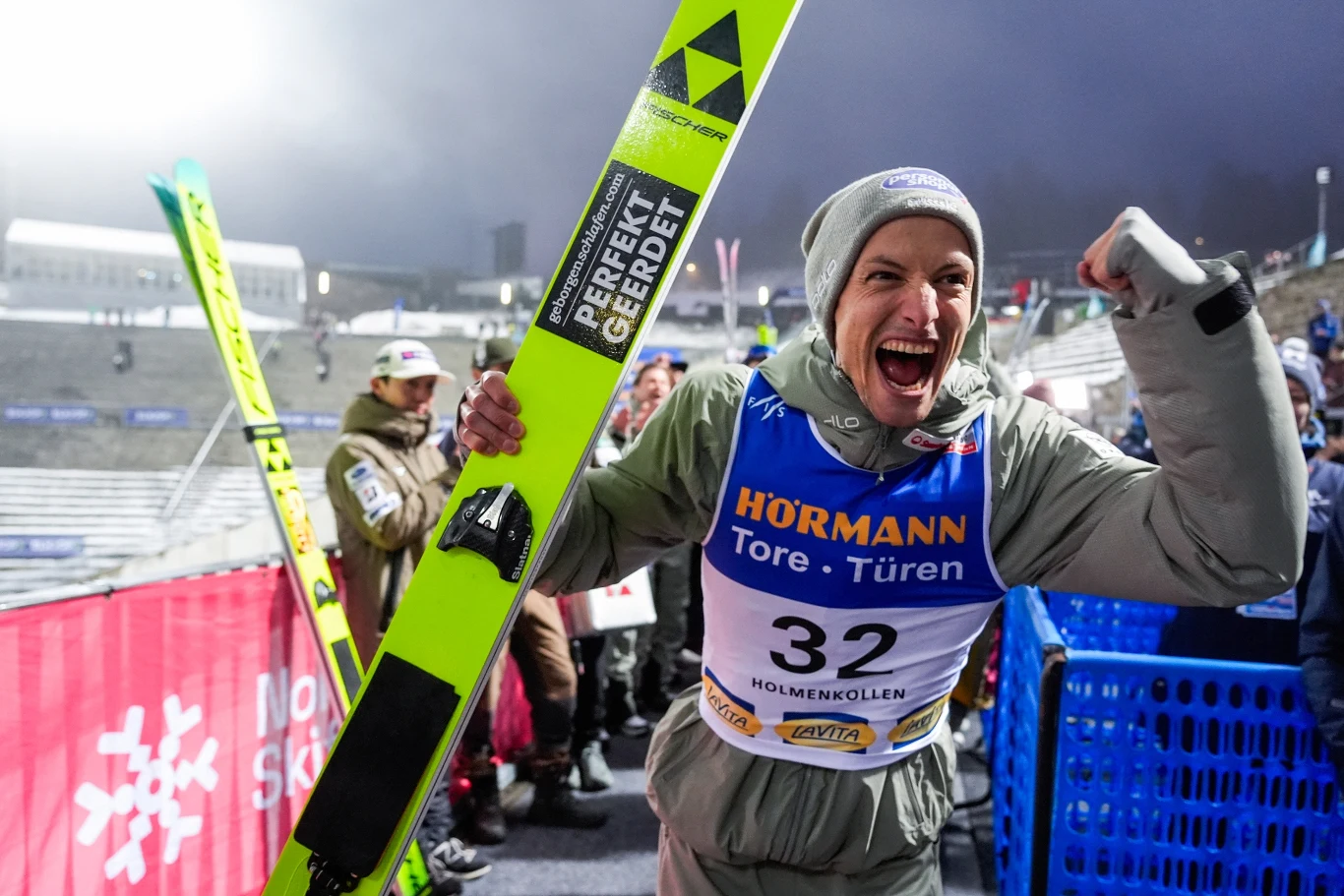 szczęśliwy skoczek narciarski w sportowym kombinezonie i czapce wykrzykuje z radości, ściskając pięść i trzymając narty, w tle zimowy stadion z kibicami oraz niebieskie ogrodzenie