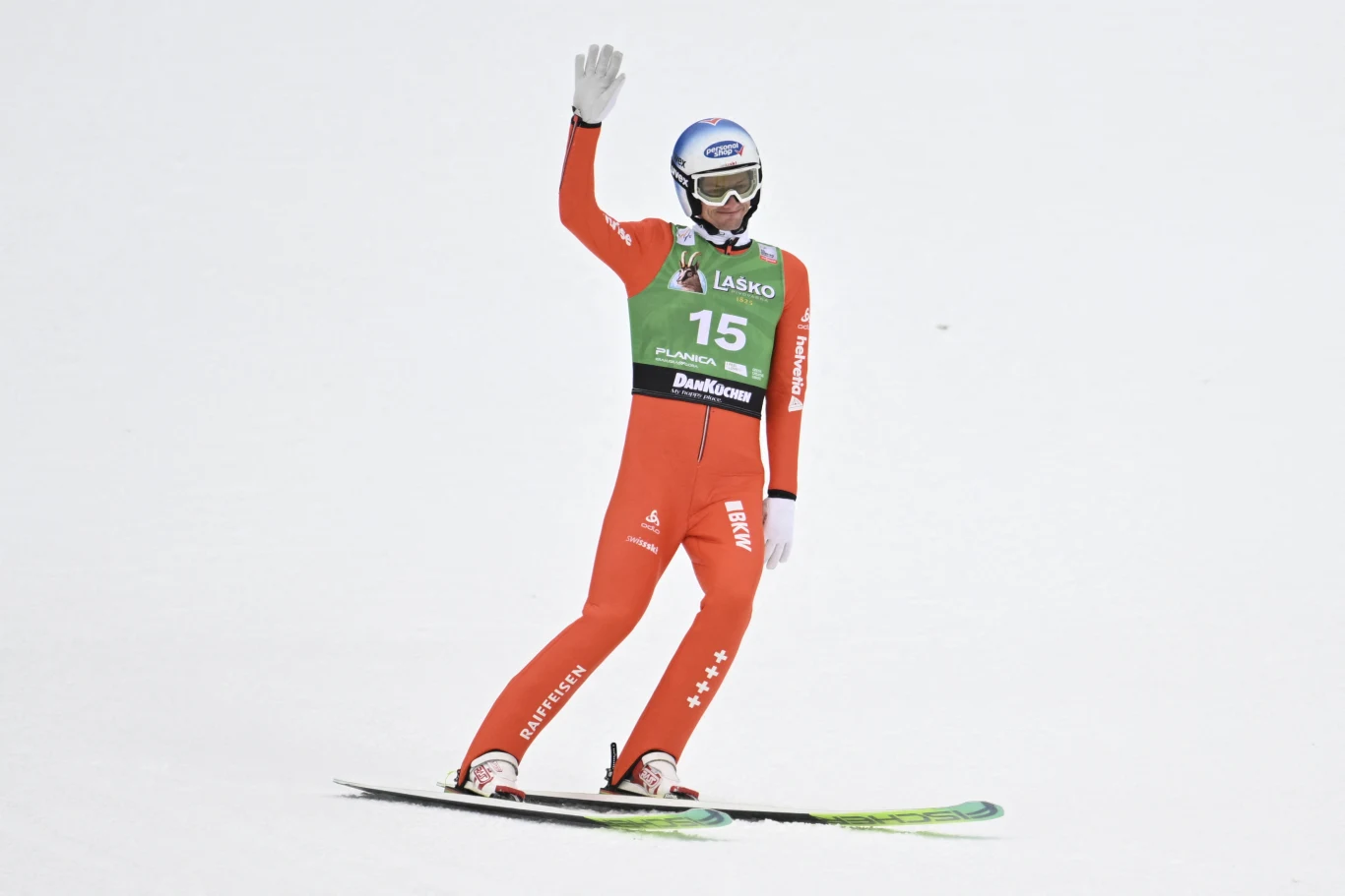 Skoczek narciarski w czerwonym kombinezonie ląduje na nartach i unosi jedną rękę w geście pozdrowienia, na sobie ma zielony plastron z numerem 15 oraz kask z goglami, w tle jednolita biała powierzchnia śniegu.