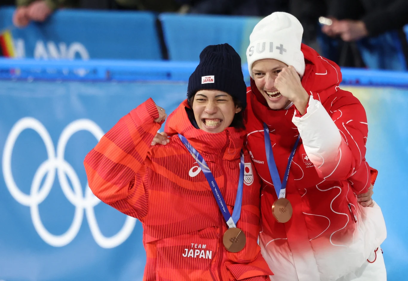 Dwie sportsmenki w olimpijskich kurtkach trzymają się i prezentują brązowe medale, uśmiechając się szeroko na tle olimpijskich kół symbolizujących igrzyska olimpijskie.