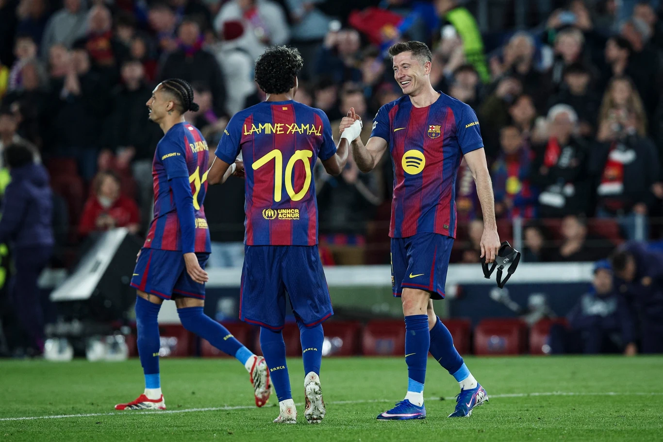 trzech piłkarzy FC Barcelony w granatowo-bordowych strojach na murawie stadionu, dwóch z nich idzie razem uśmiechniętych, jeden trzyma buty w ręku, tłum kibiców w tle