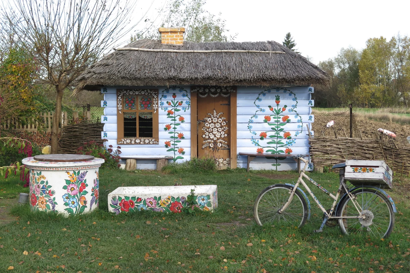 wiejska chata z drewnianych bali pokryta strzechą, dekorowana kolorowymi, ludowymi malunkami kwiatów na ścianach i okiennicach; przed domem znajduje się studnia, ławka i rower, wszystkie również ozdobione kwiatowymi motywami, w tle wiejski płot i drzewa
