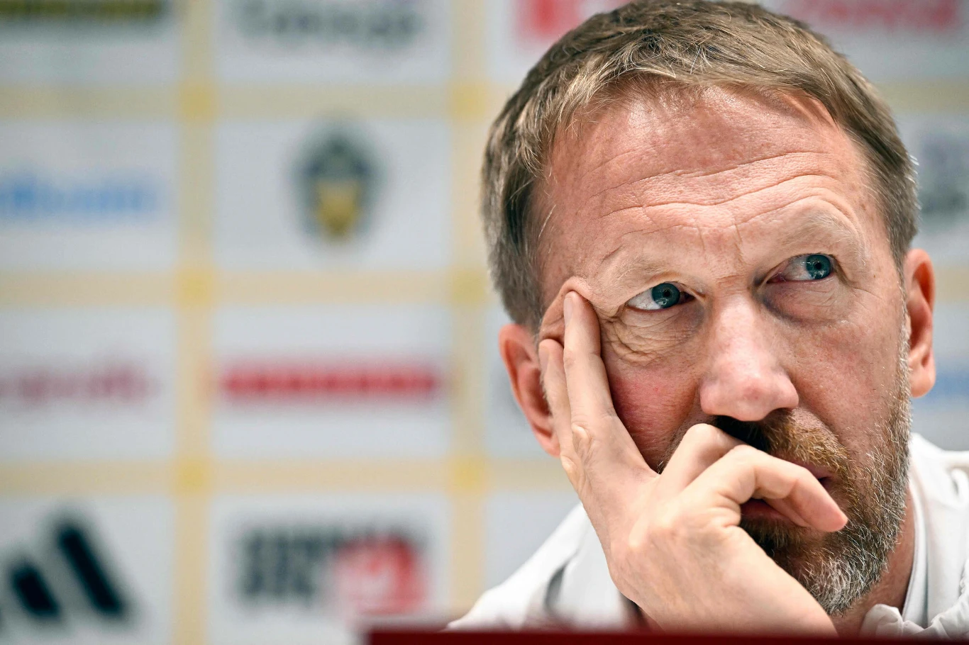 mężczyzna w średnim wieku z krótkimi, jasnobrązowymi włosami opiera głowę na dłoni i patrzy zamyślony w bok; w tle rozmazane logo i napisy typowe dla konferencji prasowej