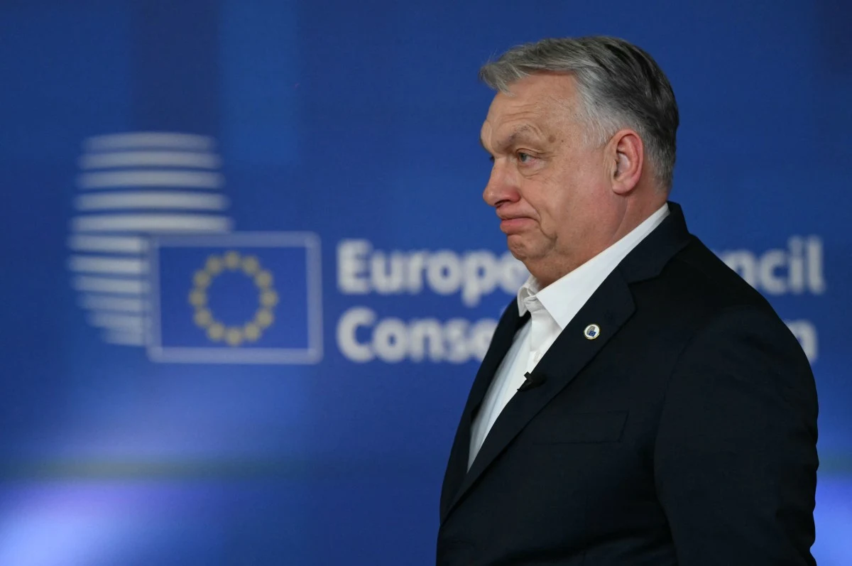 Mężczyzna w garniturze z poważnym wyrazem twarzy stoi na tle niebieskiej tablicy z logo Unii Europejskiej i napisem 'European Council' oraz okrągłym emblematem z gwiazdami.