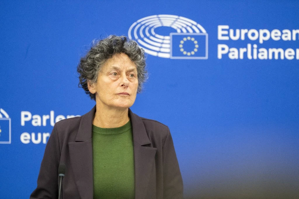 kobieta o kręconych, krótkich włosach w zielonym swetrze i ciemnej marynarce stoi przed niebieską ścianką z logo i napisem Parlamentu Europejskiego