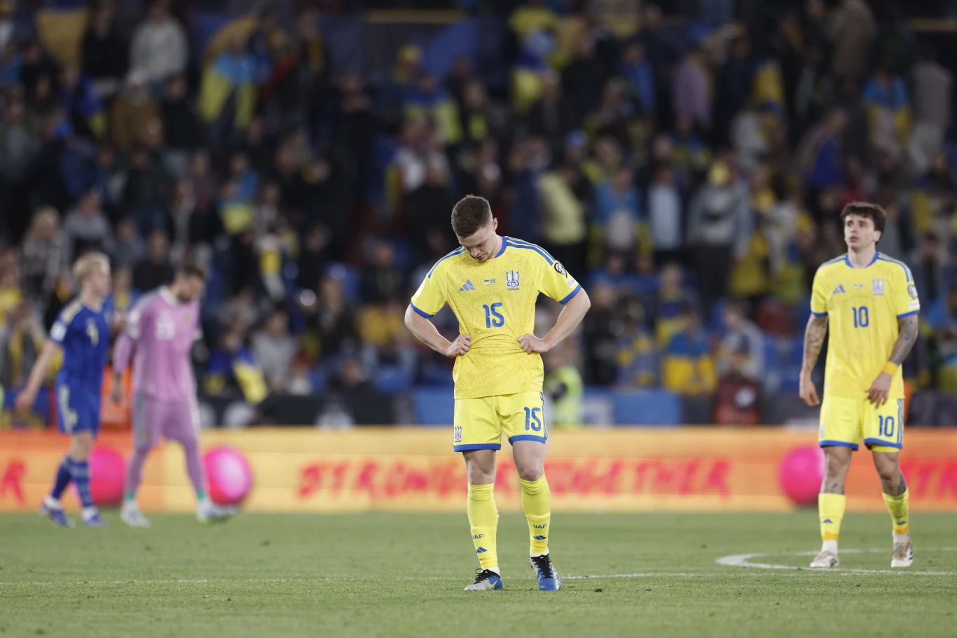 piłkarz w żółtym stroju z numerem 15 stoi ze spuszczoną głową na środku murawy, w tle widoczni są inni zawodnicy w żółtych i niebieskich strojach oraz rozmyty tłum kibiców na trybunach