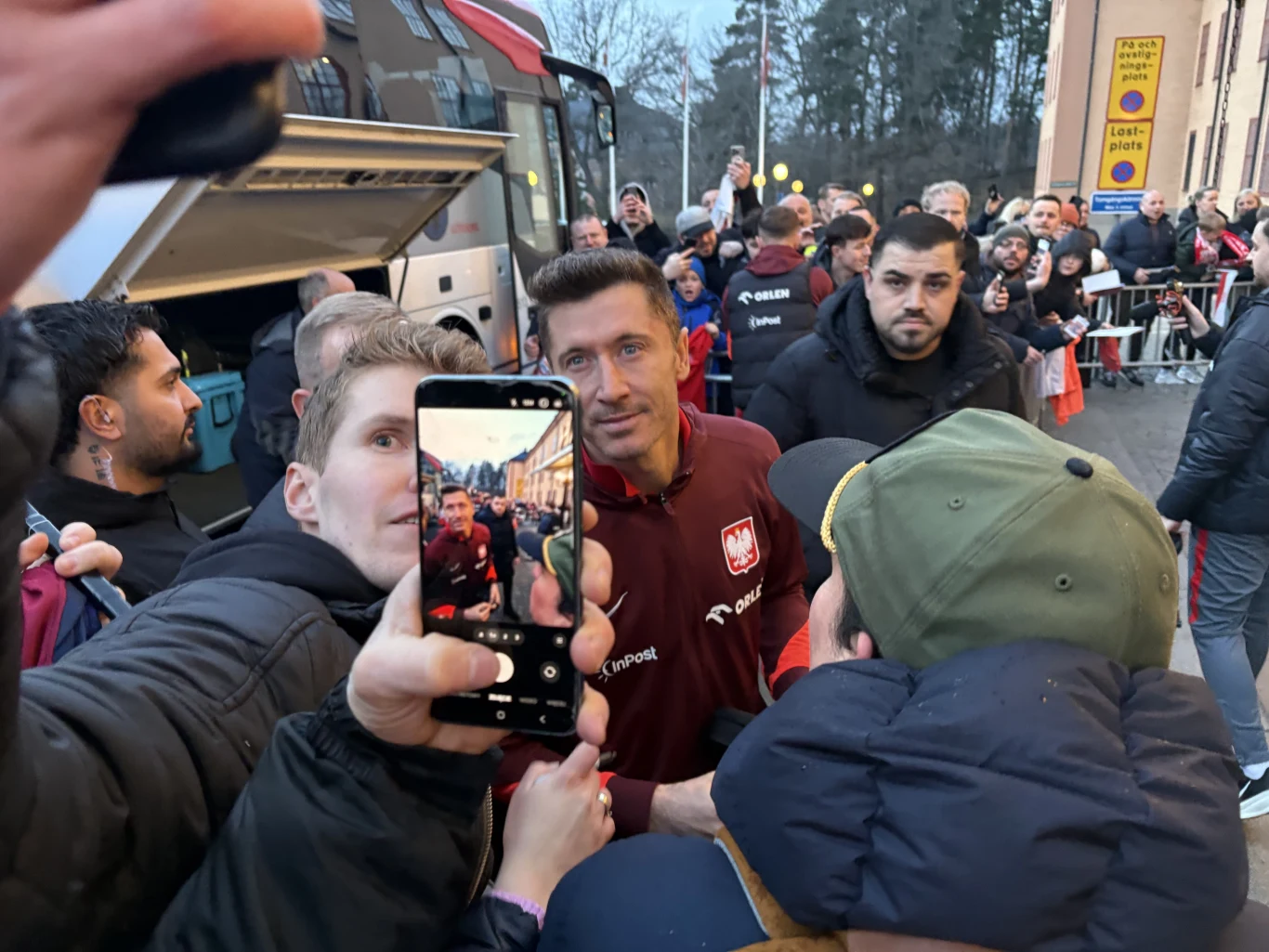 Robert Lewandowski w czerwonej kurtce reprezentacji Polski pozuje do selfie z kibicami, otoczony przez fanów i ochronę, w tle autokar oraz tłum osób oczekujących za barierkami.