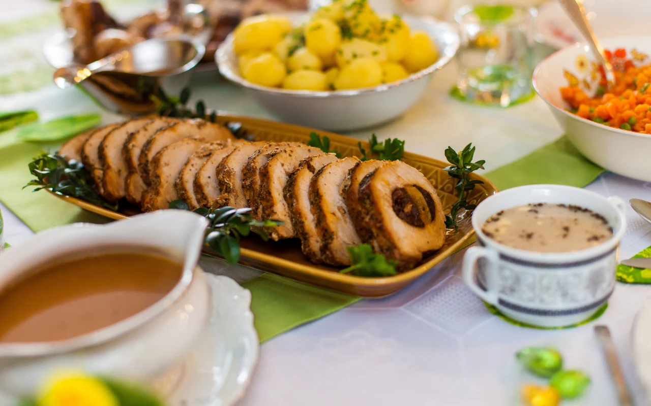 Plasterki pieczonego mięsa z ziołami ułożone na półmisku, w otoczeniu ziemniaków z koperkiem, filiżanki z sosem oraz miski z jarzynami w tle.