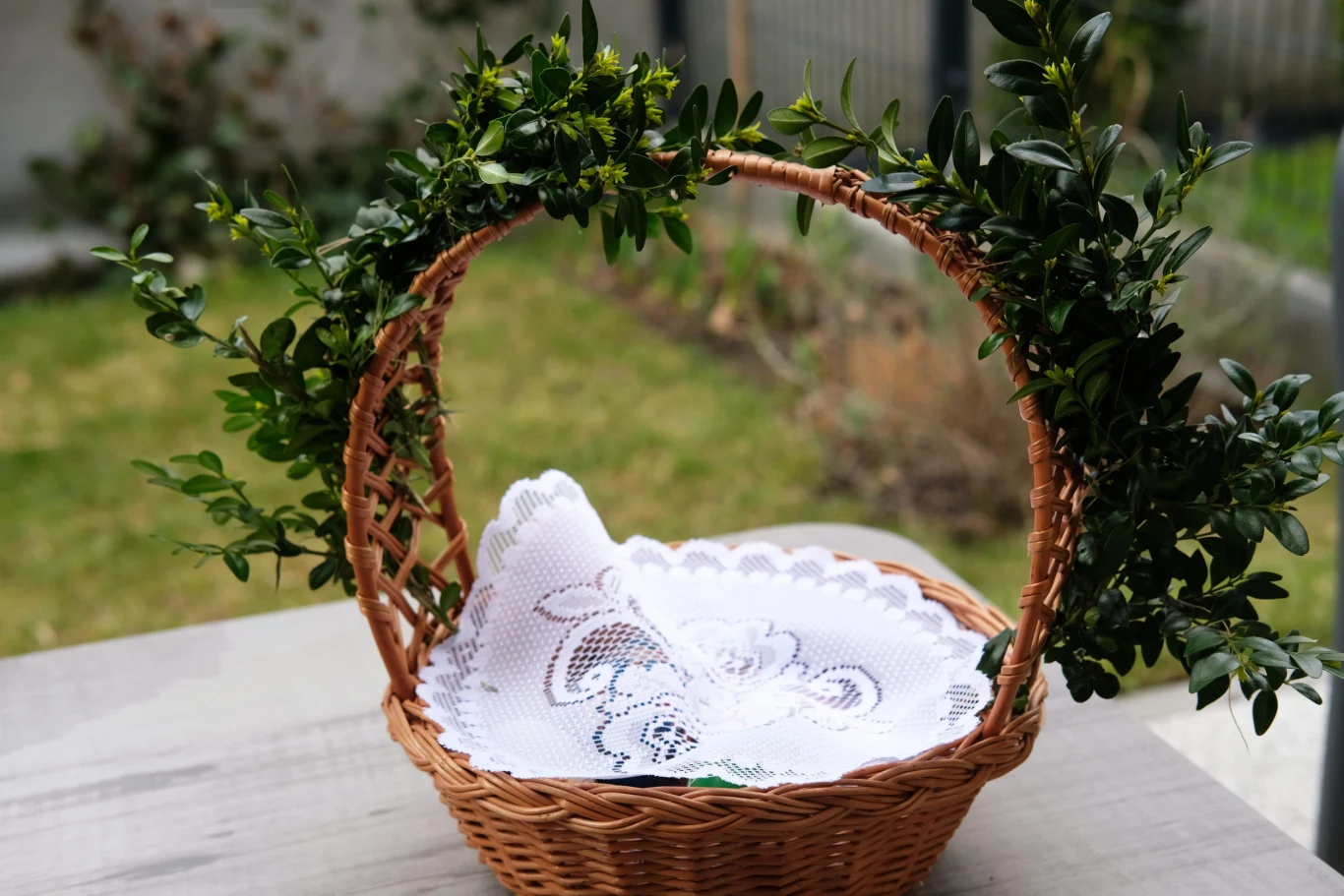 wiklinowy koszyk ozdobiony zielonym bukszpanem na uchwycie, w środku biała, haftowana serwetka