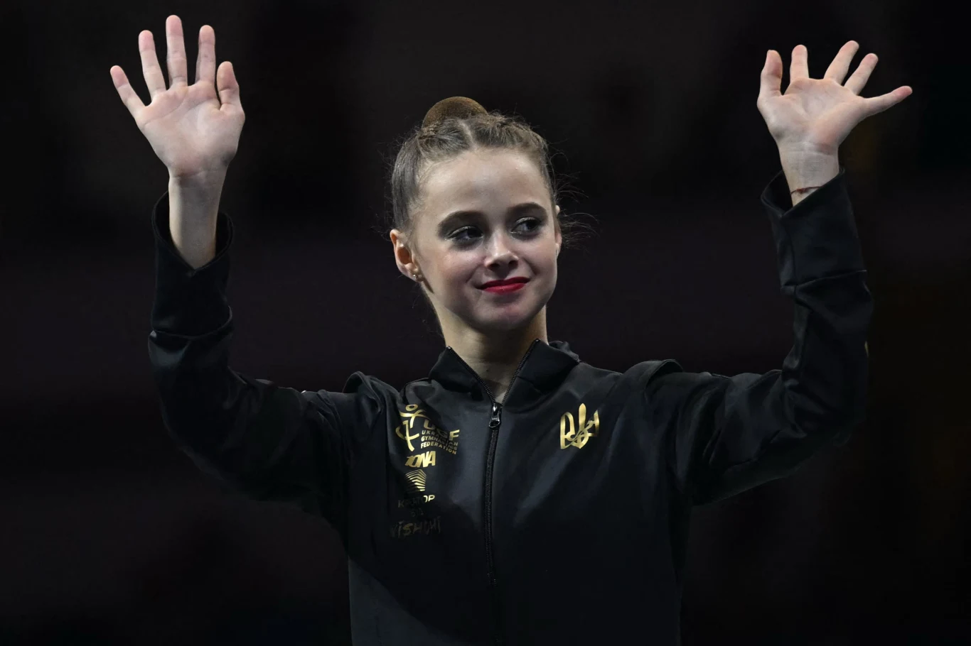 młoda kobieta w czarnej sportowej kurtce z podniesionymi rękami, uśmiechnięta, z włosami upiętymi w kok, patrzy przed siebie na tle ciemnego otoczenia