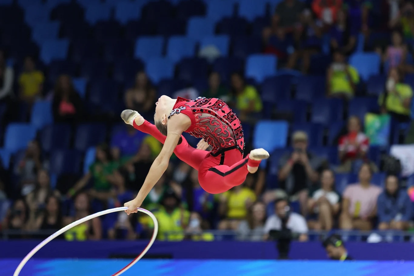 gimnastyczka w efektownym skoku z obręczą, ubrana w czerwony strój z czarnymi wzorami, dynamiczny ruch podkreślony przez wygiętą sylwetkę na tle rozmytej widowni