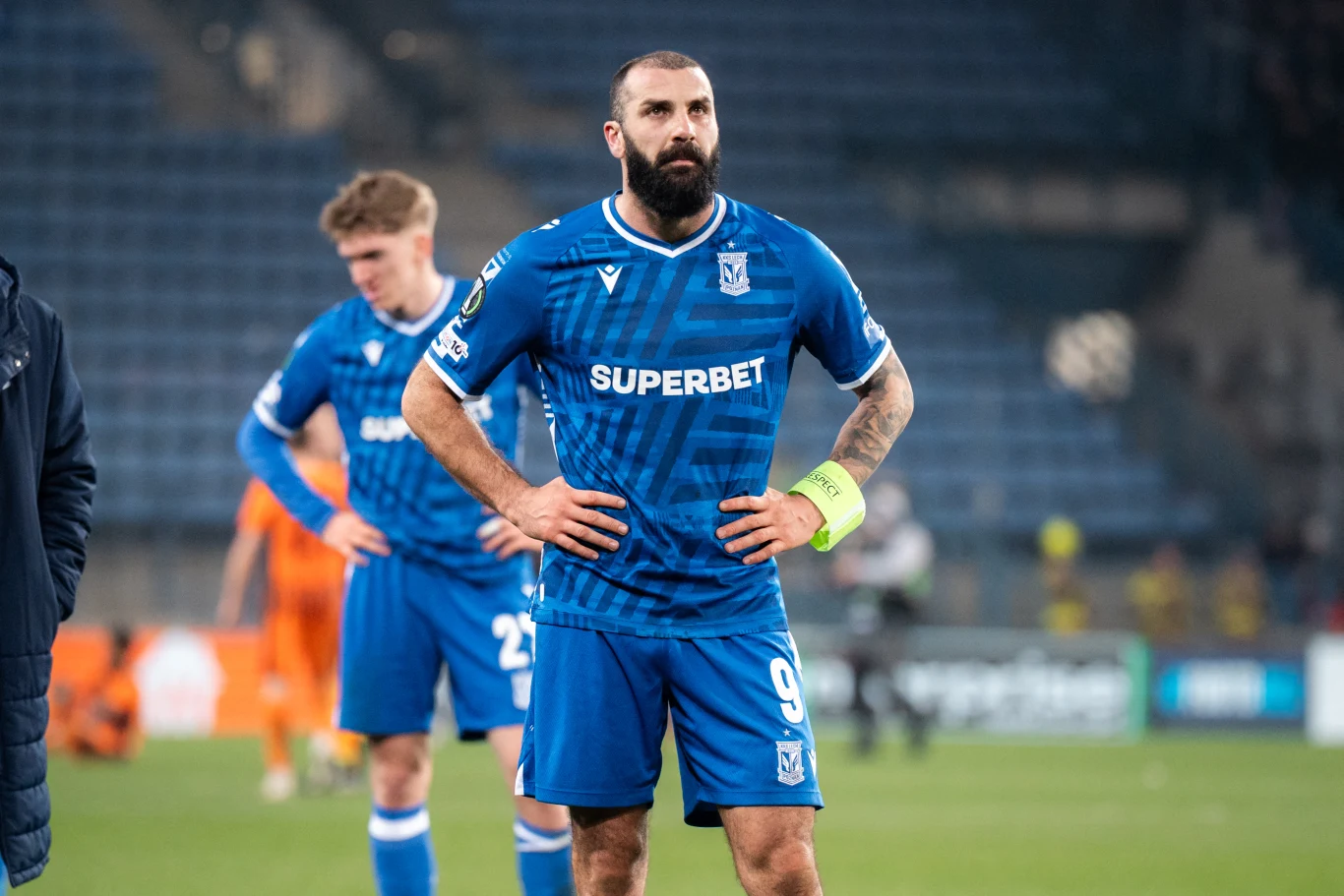 piłkarz w niebieskim stroju Lecha Poznań stoi z rękami na biodrach na murawie stadionu, na lewym ramieniu opaska kapitana, w tle inny zawodnik oraz rozmyta publiczność