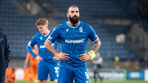 piłkarz w niebieskim stroju Lecha Poznań stoi z rękami na biodrach na murawie stadionu, na lewym ramieniu opaska kapitana, w tle inny zawodnik oraz rozmyta publiczność