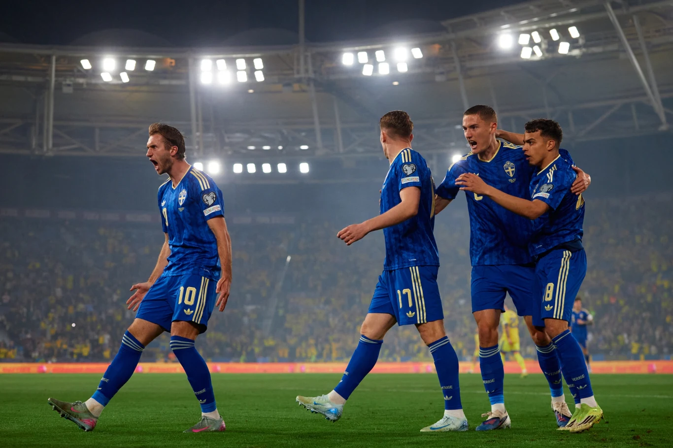 czterech piłkarzy w niebieskich strojach świętuje zdobycie gola na stadionie, na pierwszym planie jeden z graczy wyraża ogromną radość, pozostali podążają za nim z wyciągniętymi rękami, w tle rozświetlone trybuny i murawa boiska