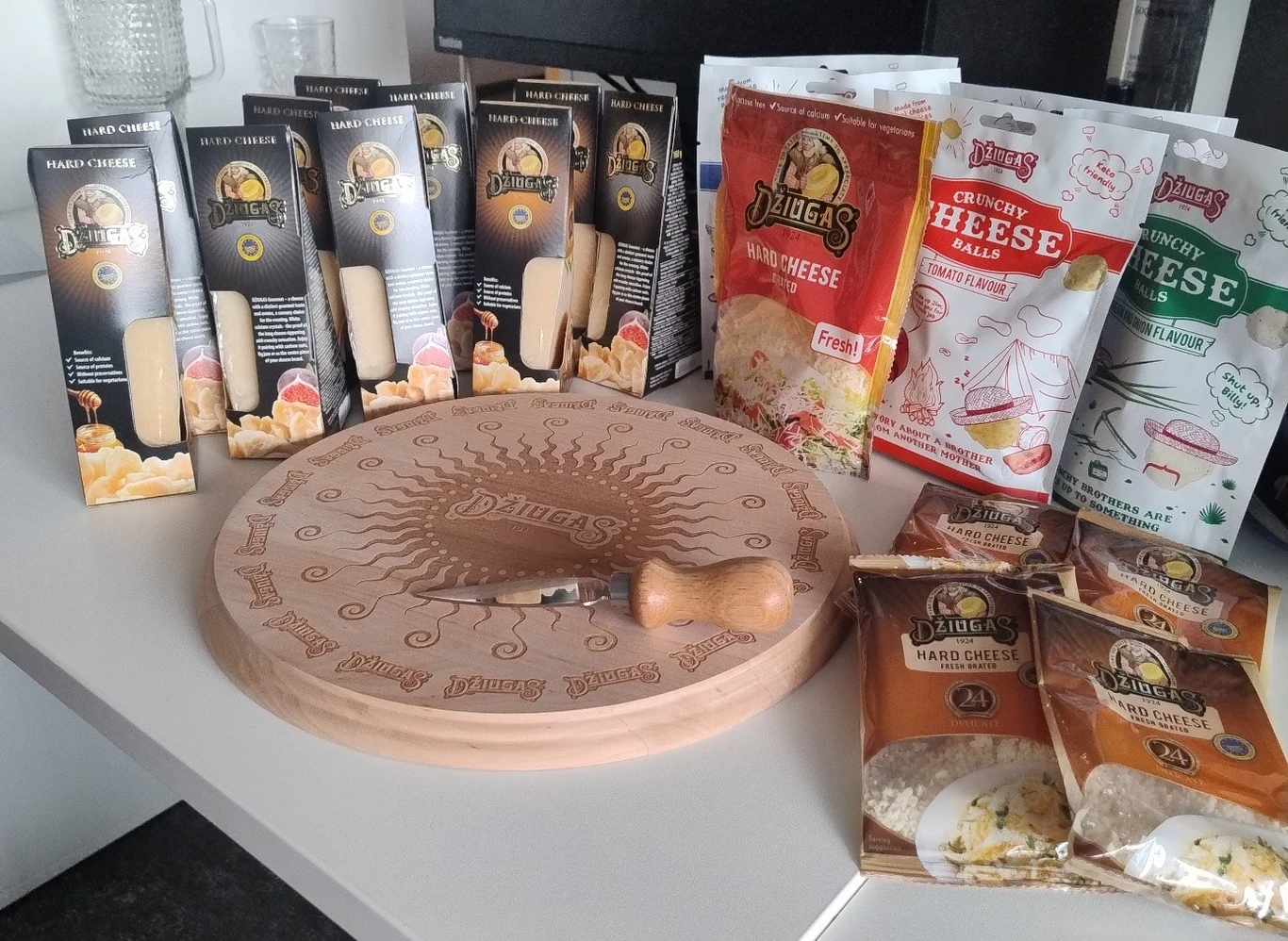 Zestaw produktów marki Džiugas obejmujący różne rodzaje twardych serów w opakowaniach, serowe przekąski i wiórki serowe, ułożone wokół ozdobnej drewnianej deski do sera z grawerem oraz specjalnym nożem do krojenia.