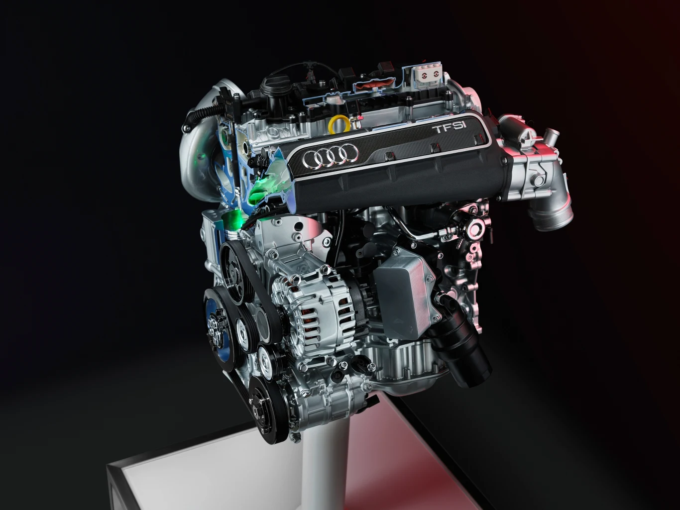silnik samochodowy Audi TFSI z widocznymi elementami konstrukcyjnymi, takimi jak turbosprężarka, alternator, przewody oraz szczegóły oznaczone logo marki i napisem TFSI, ustawiony na białym postumencie na ciemnym tle