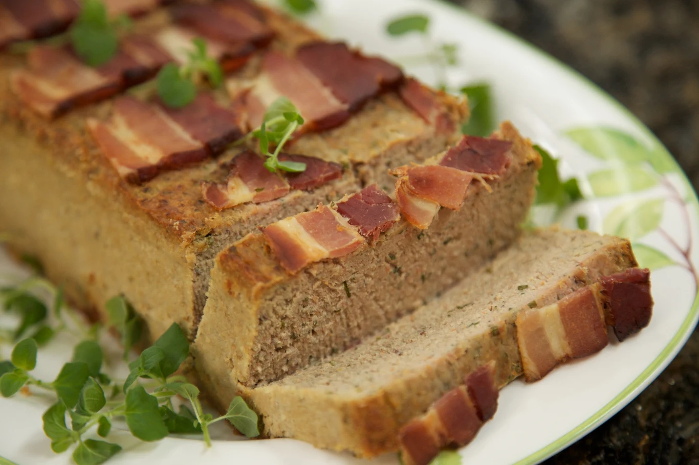 pasztet mięsny udekorowany plastrami boczku, pokrojony na grube kromki i podany na talerzu ozdobionym świeżymi ziołami