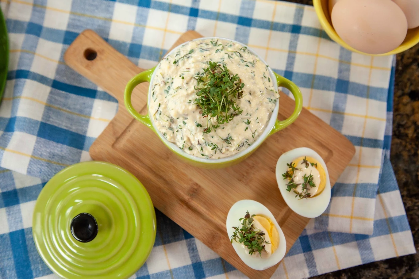 Pasta jajeczna z dużą ilością ziół w zielonym naczyniu na drewnianej desce, obok połówki ugotowanego jajka z rzeżuchą, wszystko ustawione na kraciastej ściereczce w odcieniach niebieskiego, żółtego i bieli.