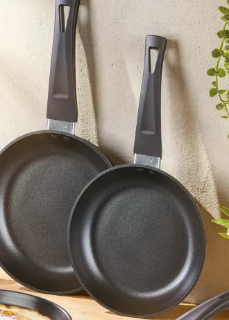 dwie czarne patelnie z nienagrzewającymi się rączkami opierającymi się o jasną ścianę, z lewej strony widoczny fragment kolejnej patelni, po prawej roślina doniczkowa