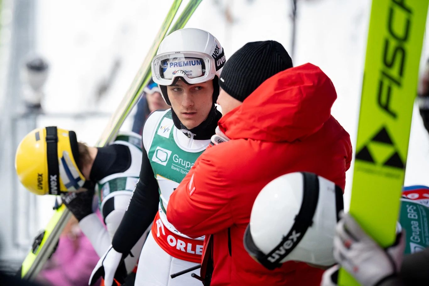 skoczek narciarski w białym kombinezonie i kasku rozmawia z trenerem ubranym w czerwoną kurtkę, obok widoczne są inni zawodnicy w sportowych strojach i narty z logo Fischer