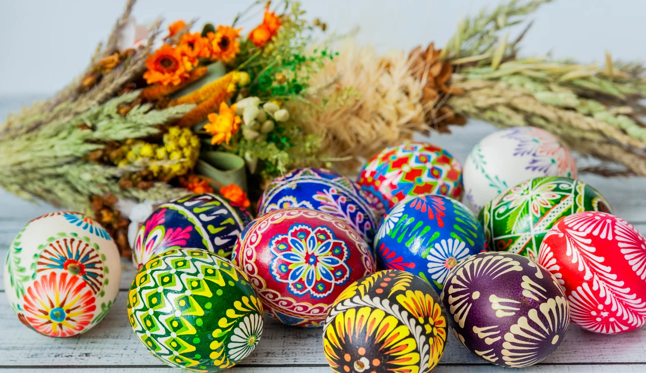 Jak składać piękne życzenia wielkanocne? Kolorowe, ręcznie malowane pisanki z ludowymi wzorami na jasnym tle, obok bukiet suszonych kwiatów i zbóż